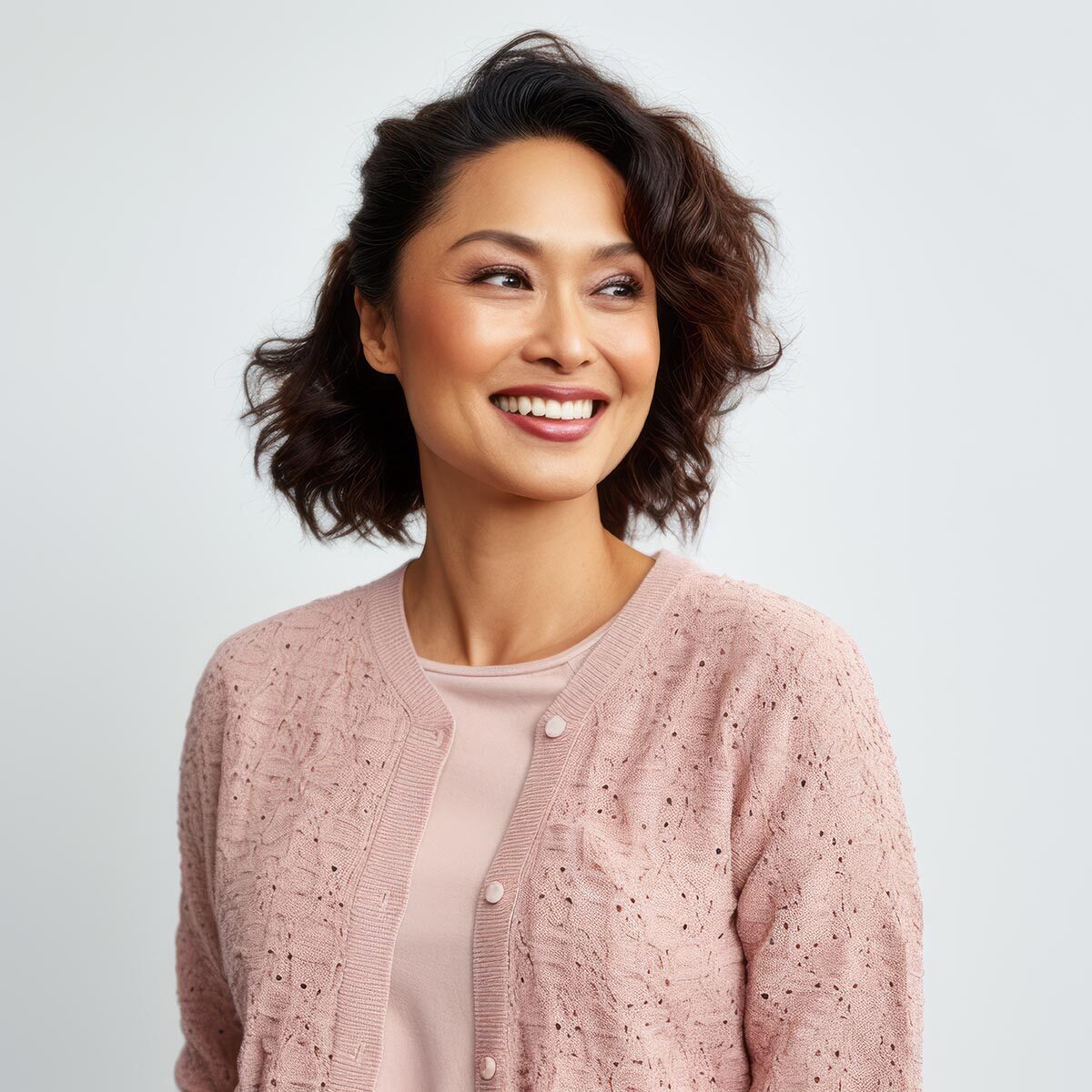 Smiling woman with short wavy hair wearing a pink cardigan on a light background.