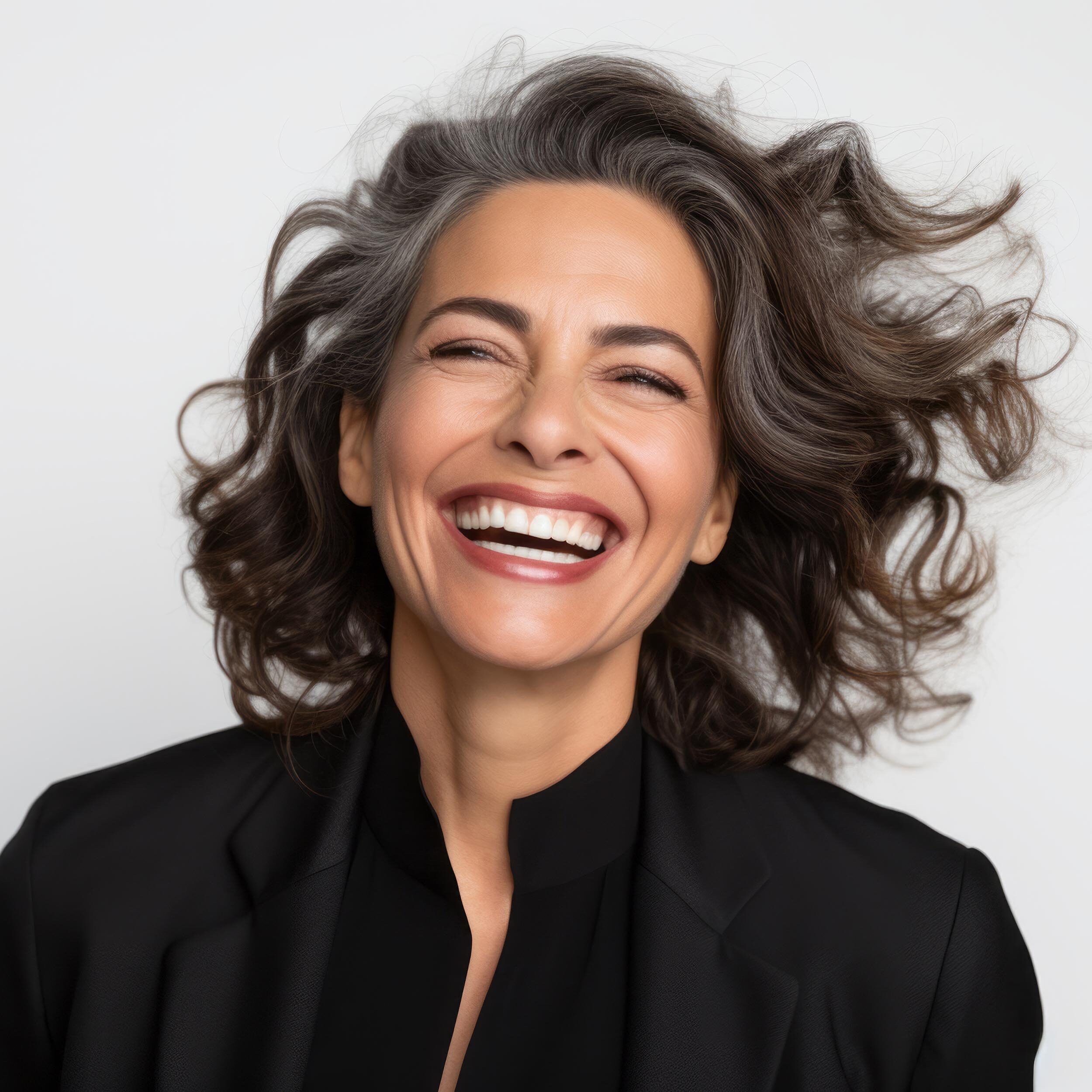 Woman with curly hair laughing heartily in a black blazer on a white background.