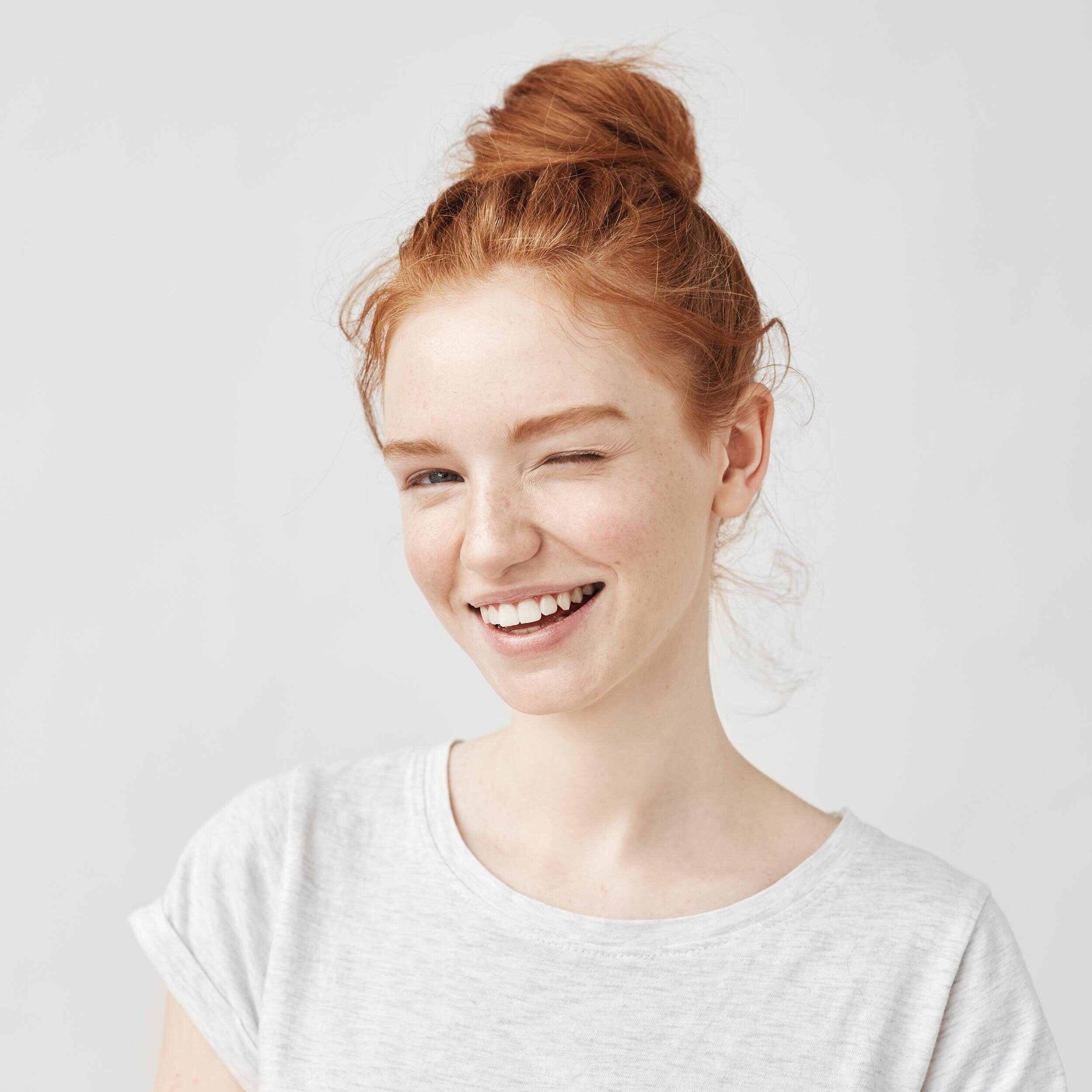 Young woman with red hair tied in a bun, winking and smiling, wearing a light gray t-shirt on a white background.