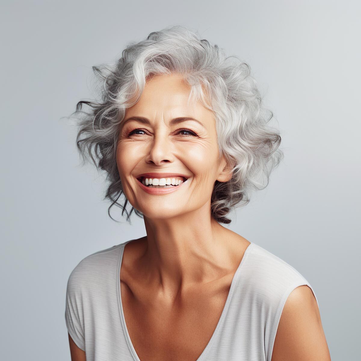 Senior woman with curly gray hair and a radiant smile, wearing a white top.
